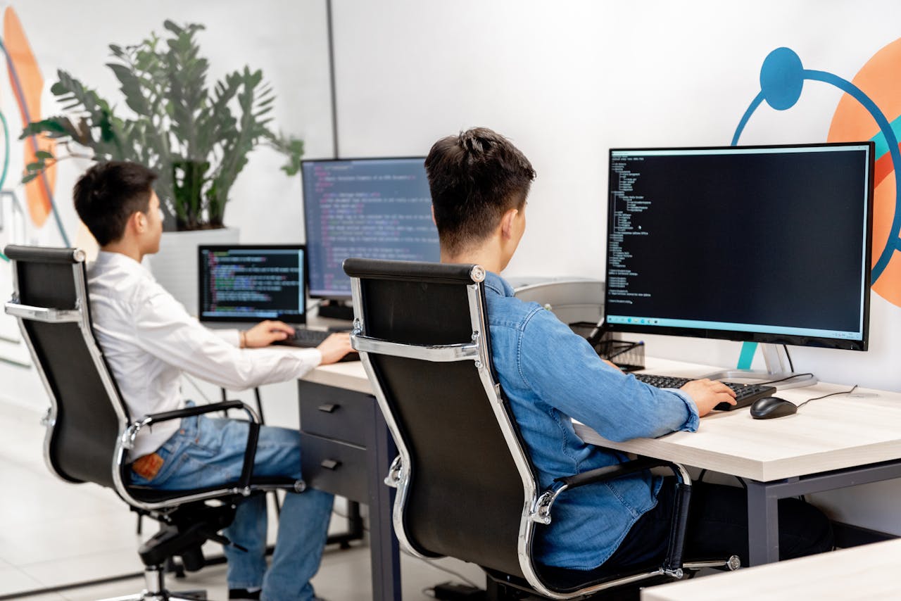 our-services-2 Two male developers at desks programming in a modern office workspace with large monitors.